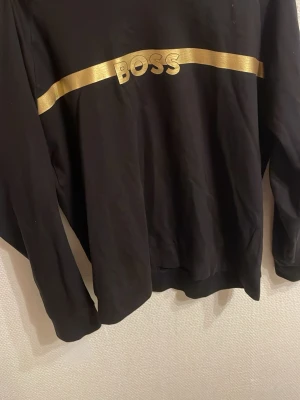 Svart sweatshirt från BOSS med guldtryck - Säljer en svart sweatshirt från BOSS Homewear i storlek L. Tröjan har ett fett guldfärgat band och BOSS-logga över bröstet. Klassisk rund halsringning och långa ärmar. Perfekt för dig som vill ha en clean men ändå statement look.