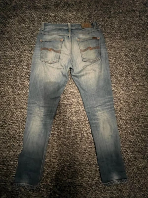 Ljusblå jeans från Nudie Jeans, Grim Tim - Säljer ett lite par shysstare ljusblå slim jeans från Nudie Jeans med klassiska orange sömmar och snygga slitningar. Jeansen har fem fickor, bälteshällor och en smal passform som sitter tajt längs benen. Perfekta för dig som gillar en clean och modern look.