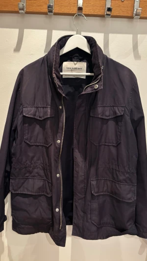 Mörkblå fieldjacka från These Glory Days - Stilren mörkblå jacka från These Glory Days med fyra stora fickor framtill, hög krage och dragkedja med tryckknappar. Jackan har en rak passform och är tillverkad i ett lätt, slitstarkt material som passar perfekt till vardagslooken.