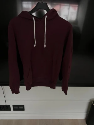 Vinröd hoodie från Pier One - Säljer en vinröd hoodie från Pier One med klassisk huva och vita dragsnören. Hoodien har en stor magficka framtill och ribbade muddar vid ärmar och nederkant. Perfekt för en chill och avslappnad stil. Materialet känns mjukt och bekvämt.