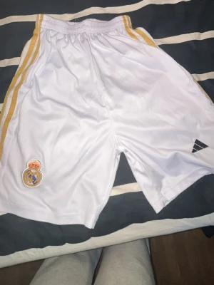 Vita Real Madrid shorts från Adidas - Snygga vita shorts från Adidas med Real Madrid-logga och guldiga ränder längs sidorna. Perfekta för fotboll eller träning, med elastisk midja och sportig design.