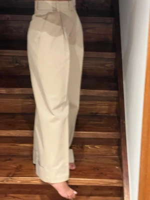 Beiga vida chinos med hög midja - Snygga beiga chinos med hög midja och raka, vida ben. Klassisk look med pressveck och bred fåll nedtill. Perfekta för en clean och stilren outfit. Tillverkade i mjuk bomull som känns skön mot huden. 