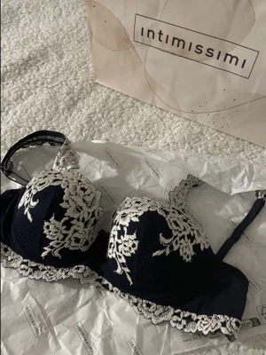 Mörkblå spets-bh från Intimissimi - Snygg mörkblå bh från Intimissimi med vita spetsdetaljer och blommönster. Bh:n har vadderade kupor och justerbara axelband. Perfekt för dig som gillar en elegant och feminin stil.