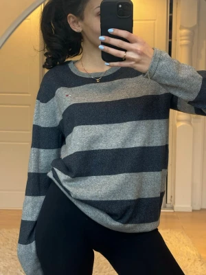 Randig stickad tröja Tommy Hilfiger - Mysig stickad tröja från Tommy Hilfiger med breda mörkgrå och ljusgrå ränder. Tröjan har rund halsringning och en liten broderad logga på bröstet. Perfekt för en avslappnad och stilren look.