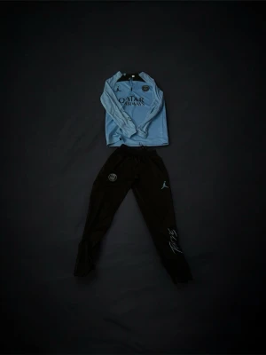 PSG träningsset Jordan x Nike - Snyggt PSG träningsset med ljusblå långärmad tröja och svarta byxor. Tröjan har Qatar Airways-logga och PSG-märke, byxorna har PSG-logga och Jordan-logga samt tryck i ljusblått. Perfekt för fotbollsträning eller chill.Byxorna har även ett litet hål i sig. (Se bild) storleken är 164