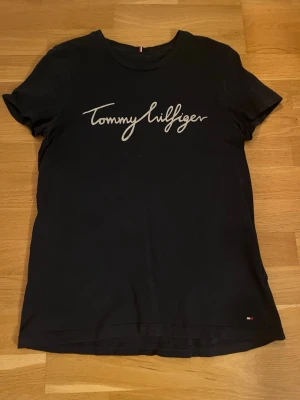 Svart Tommy Hilfiger t-shirt - Svart t-shirt från Tommy Hilfiger med vit logga i skrivstil över bröstet. Klassisk rund halsringning och korta ärmar. Liten flaggdetalj nere vid fållen. Perfekt till jeans eller shorts.