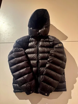 Svart dunjacka med huva från Bvaldi - Snygg svart dunjacka från Bvaldi med huva och dragkedja framtill. Jackan har quiltad design och Bvaldi-logga på ärmen. Perfekt för kalla dagar och har en modern, puffig look som är riktigt trendig just nu. Bara skriva vid funderingar🙌 Jackan kostar 3000 ny!