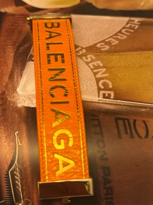 Orange läderarmband från Balenciaga - Snyggt orange armband i läder från Balenciaga med guldiga metalldetaljer och logotyp i guldtext på framsidan. Armbandet har en bred design och känns riktigt lyxigt och trendigt. Perfekt accessoar för att lyfta din outfit.