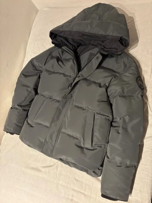 Mörkgrå pufferjacka från Canada Goose - Snygg mörkgrå pufferjacka från Canada Goose med huva och diskret logga på ärmen. Jackan har dragkedja och knappar framtill samt två stora fickor. Perfekt för kalla dagar och riktigt skön passform.