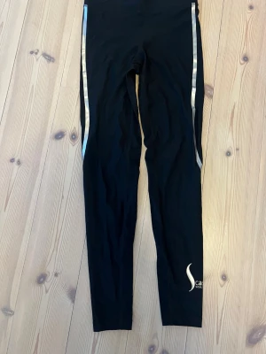 Svarta träningstights från Casall - Snygga svarta träningstights från Casall med silvriga ränder längs benen. Deras sculpture modell. .