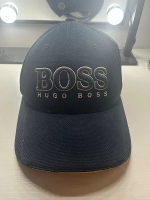 Marinblå keps från Hugo Boss - Snygg marinblå keps från Hugo Boss med stor vit/blå broderad BOSS-logga framtill och texten HUGO BOSS under. Klassisk böjd skärm och justerbar baksida. Tillverkad i ett tåligt och ventilerande material som passar perfekt till streetwear och casual outfits. PRIS kan diskuteras, finns fler bilder om man vill ha.
