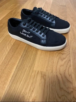 Svarta Saint Laurent sneakers - Svarta sneakers från Saint Laurent med vit sula och handskriven logga på sidan. Skorna har snörning och är tillverkade i en mix av textil och skinn för en clean och stilren look. Perfekta för dig som gillar minimalistisk design.
