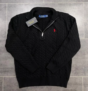 Svart kabelstickad tröja Polo Ralph Lauren - Kabelstickad svart tröja från Polo Ralph Lauren med halv dragkedja och röd broderad logga på bröstet. Tröjan har ribbade muddar och krage, samt klassisk passform. Perfekt för dig som gillar stilrena och tidlösa plagg.