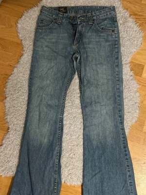Blå bootcut jeans från Lee - Säljer ett par klassiska blå bootcut jeans från Lee. Jeansen har fem fickor, normal passform och är tillverkade i starkt denimtyg av bomull. De har en ljus tvätt och Lee-logga på bakfickan. Perfekta för en avslappnad och trendig look.