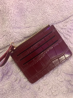 Vinröd plånbok i croc-mönstrat skinn - Snygg vinröd plånbok med croc-mönster och guldfärgad dragkedja. Plånboken har flera kortfack på framsidan och en handledsrem för enkel användning. Perfekt för att hålla koll på kort och kontanter i stil.