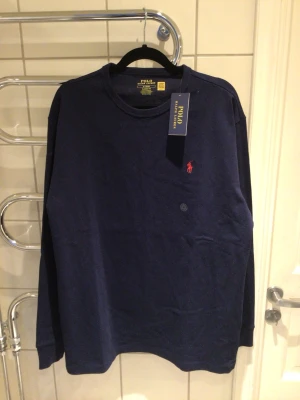Marinblå långärmad tröja Polo Ralph Lauren - Snygg marinblå oanvänd långärmad tröja från Polo Ralph Lauren i klassisk passform. Tröjan har rund halsringning och den ikoniska röda logotypen broderad på bröstet. Tillverkad i mjuk bomull som känns skön mot huden.