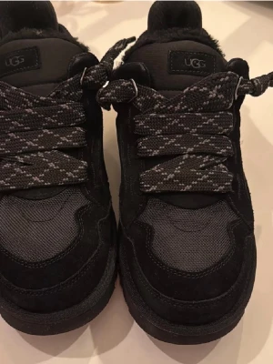 Svarta sneakers från UGG - Säljer ett par svarta sneakers från UGG med breda, mönstrade skosnören och robust sula. Skorna har en kombination av mocka och textilmaterial samt UGG-logga på plösen och sulan. Perfekta för dig som gillar en chunky look.