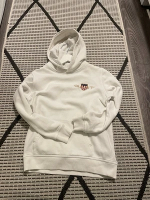 Vit hoodie från GANT med logga - Snygg vit hoodie från GANT med broderad logga och sköld på bröstet. Klassisk huva och känguruficka framtill. Tillverkad i mjukt bomullsmaterial som känns skönt mot huden. Perfekt för en avslappnad och stilren look.
