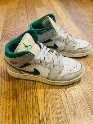 Nike Air Jordan 1 Mid vit/grön - Nike Air Jordan 1 Mid sneakers i vitt och grönt med klassisk Jordan-logga på plösen och gröna detaljer. Skorna har snörning, perforerad tå och en bekväm vadderad krage. Perfekta för dig som gillar streetstyle och ikoniska sneakers.