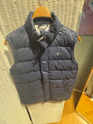 Mörkblå dunväst från GANT - Snygg mörkblå dunväst från GANT med hög krage och diskret logga på bröstet. Västen har quiltad design, dragkedja framtill och två fickor. Perfekt för lager på lager och ger en sportig, clean look.