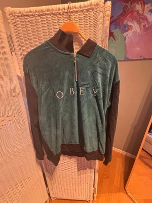 Grön ziptröja från OBEY - Mjuk grön ziptröja från OBEY med svart krage och svarta ärmar. Tröjan har halv dragkedja framtill och OBEY-loggan broderad över bröstet. Perfekt för en chill och avslappnad stil.