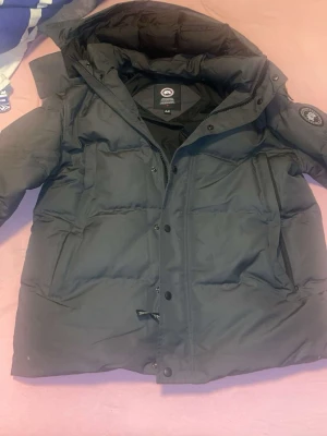 Svart dunjacka från Canada Goose XL - Säljer en svart dunjacka från Canada Goose i storlek XL. Jackan har huva, dragkedja och knappar framtill samt två sidofickor med dragkedja. Perfekt för kalla dagar och riktigt varm med klassisk patch på ärmen.