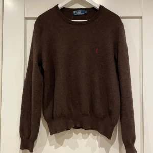 Brun merinoull från Polo Ralph Lauren - Mysig brun tröja från Polo Ralph Lauren i merinoull med rund halsringning och diskret röd broderad logga på bröstet. Klassisk passform och ribbade muddar vid ärmslut och nederkant. Perfekt för lager på lager under höst och vinter.