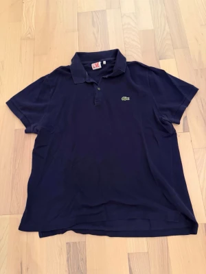Mörkblå pikétröja/tshirt från Lacoste  - Klassisk mörkblå pikétröja från Lacoste med korta ärmar och krage. Tröjan har två knappar framtill och den ikoniska gröna krokodilloggan på bröstet. Tillverkad i mjuk bomull som känns skön mot huden. Storleken är 6 men passar mig som har storlek L på kläder