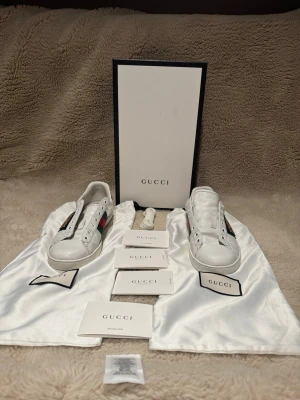 Gucci vita sneakers med gröna och röda detaljer - Säljer ett par klassiska vita sneakers från Gucci med gröna och röda ränder på sidan. Skorna har rund tå, snörning och är tillverkade i skinn. Medföljer originalkartong, dustbags, extra skosnören och äkthetskort.