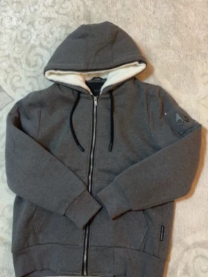 Moose Knuckles bunny zip  - Snygg grå hoodie med dragkedja från Moose Knuckles. Hoodien har en mjuk vit fuskpälsfodrad huva, svarta dragsnören och två fickor framtill. Detalj med logotyp på ärmen och ribbade muddar vid ärmslut och nederkant. Perfekt för en chill och stilren look.