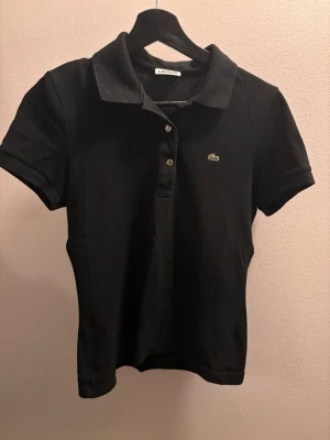 Svart pikétröja/t-shirt från Lacoste dam 38 - Klassisk svart pikétröja från Lacoste med korta ärmar och krage. Tröjan har två knappar framtill och den ikoniska gröna krokodilloggan på bröstet. Litegrann ”urtvättad” i färgen, men annars i mycket fint skick och mycket lite använd.