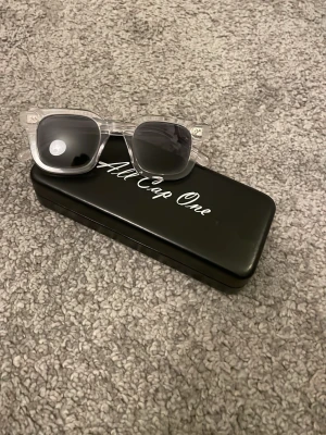 Transparenta solglasögon All Cap One - Snygga solglasögon från All Cap One med transparent båge och mörka glas. Klassisk fyrkantig form som passar till många stilar. Kommer med ett svart fodral med vit logga. Perfekt accessoar för soliga dagar.