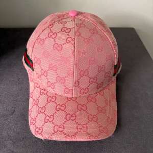 Rosa Gucci keps med GG-mönster - Exklusiv rosa keps från Gucci med ikoniskt GG-monogram över hela ytan. Kepsen har gröna och röda ränder på sidan och justerbar rem baktill i rosa läder. Tillverkad i Italien i bomullsmaterial, perfekt för dig som vill sticka ut med en lyxig accessoar.
