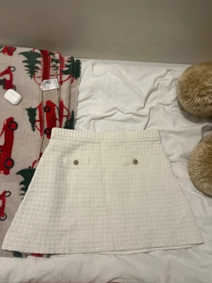 Vit kortkjol från H&M i stickad look - Supersöt vit kortkjol från H&M med ribbad midja och två dekorativa knappar framtill. Kjolen har en stickad struktur och är lätt utställd för en chill siluett. Perfekt att styla med en enkel topp eller oversized tröja.