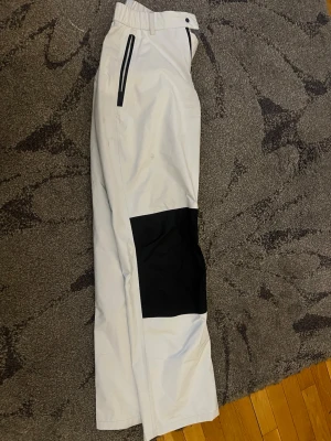 Vita täckbyxor Sail Racing - Snygga vita täckbyxor från Sail Racing med svarta detaljer och förstärkningar på knäna. Byxorna har dragkedjeförsedda fickor och logga på ena benet. Perfekta för vinterns alla äventyr och håller dig både varm och torr.