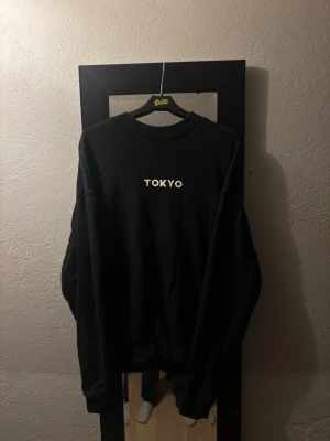 Svart sweatshirt med TOKYO-tryck - Cool svart sweatshirt med vitt TOKYO-tryck på bröstet. Tröjan har rund halsringning och ribbade muddar vid ärmslut och nederkant. Perfekt för en avslappnad streetwear-look.