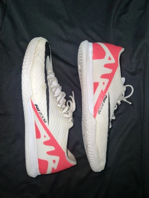 Nike vita och röda fotbollsskor - Säljer ett par vita Nike fotbollsskor med stora rosa detaljer och svart swoosh på sidan. Skorna har snörning och platt sula, perfekta för inomhusspel. Tillverkade i syntetmaterial för lätt känsla och bra grepp på planen.