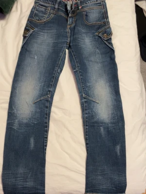 Blå jeans med stora bakfickor - Snygga blå jeans med slitningar och unika stora bakfickor. Jeansen har kontrastsömmar, knappar vid fickorna och en klassisk femficksdesign. Perfekta för dig som vill sticka ut med detaljer och cool tvätt.