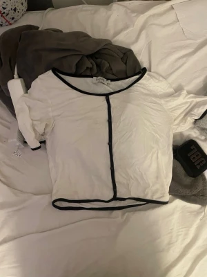 Vit topp med svarta kanter från Na-kd - Vit kortärmad topp från Na-kd med svarta kontrastkanter längs halsringning, knappslå och nederkant. T-shirten har en enkel, croppad passform och är tillverkad i mjukt bomullsmaterial. Perfekt till jeans eller kjol för en clean look.