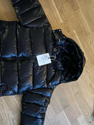 Moncler Maya - Snygg svart pufferjacka från Moncler med glansig finish och huva. Den är i storlek M=3 och e perfekt till vintern dm mig för minsta lilla fråga☺️