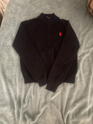 Svart stickad kofta från Polo Ralph Lauren - Svart ribbstickad kofta från Polo Ralph Lauren med dragkedja framtill och klassisk röd broderad logga på bröstet. Hög krage och långa ärmar, perfekt för lager på lager under höst och vinter. Snygg och enkel att matcha till det mesta.