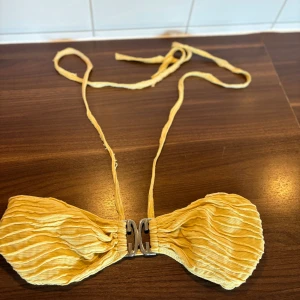 Gul bandeau bikiniöverdel med struktur - Snygg gul bandeau bikiniöverdel med vågig struktur och dekorativt metallspänne framtill. Bikinin har tunna knytband i nacken och är tillverkad i ett stretchigt material som ger en bekväm passform. Perfekt för strandhäng och soliga dagar. Bikini topp med trosan och en kjol kommer med 