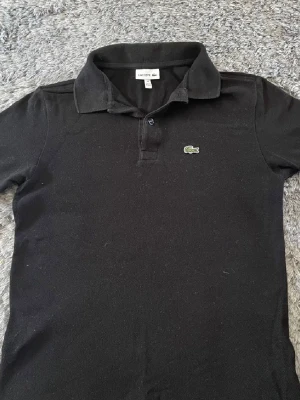 Svart pikétröja från Lacoste - Klassisk svart pikétröja från Lacoste med korta ärmar och den ikoniska gröna krokodilloggan på bröstet. Tröjan har krage och knäppning framtill. Perfekt till både jeans och kjol för en sportig och stilren look.