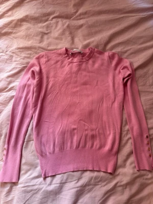 Rosa tröja - Söt rosa stickad tröja med rund hals och långa ärmar. Ärmsluten har dekorativa guldfärgade knappar och en liten slits. Tröjan har ribbade muddar vid midjan och ärmarna för en snygg passform. Perfekt för dig som gillar en feminin och stilren look.