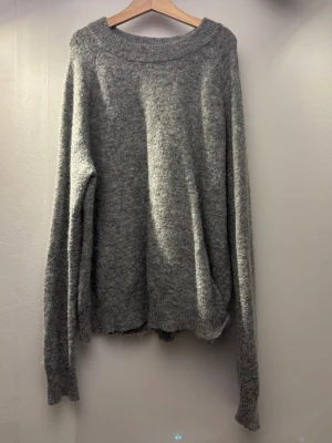 Grå stickad tröja med bred hals - Mysig grå stickad tröja med bred halsringning och långa ärmar. Tröjan har en avslappnad passform och ribbstickade muddar vid ärmslut och nederkant. Lätt att styla och har ett mycket bra skick!