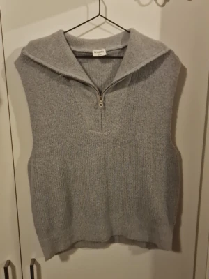 Grå stickad väst från Stories - Mysig grå stickad väst från Stories med hög krage och halv dragkedja framtill. Västen är ärmlös och har en avslappnad passform, perfekt att slänga över en skjorta eller tröja. Ribbstickad struktur ger en trendig look.