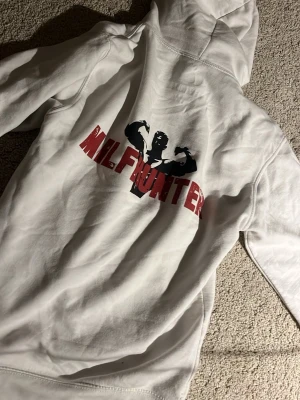 Vit hoodie MILFHUNTER - Säljer en vit hoodie med huva och ett stort tryck på ryggen där det står 'MILFHUNTER' i rött och en svart siluett av en bodybuilder. Litet hål i armhålan men syns såklart inte👍 Hoodien har klassisk passform och är tillverkad i mjukt bomullsmaterial. Använd typ 4 ggr och är eftertraktad eftersom den inte tillverkas längre. Revenite är borta och klädesplaggen går upp i värde📈. Skickas snabbt!🛫