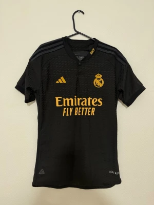 Real Madrid svart matchtröja Adidas ( inget namn ) - Snygg svart Real Madrid matchtröja från Adidas med gula detaljer och klubbmärke på bröstet. Tröjan har korta ärmar, rund hals och trycket 'Emirates Fly Better' framtill. Tillverkad i lätt och ventilerande funktionsmaterial, perfekt för fotbollsträning.