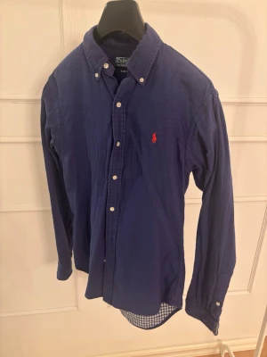 Blå skjorta från Polo Ralph Lauren - Klassisk blå skjorta från Polo Ralph Lauren med knappkrage och den ikoniska röda logobrodyr på bröstet. Skjortan har lång ärm, knappar framtill och är tillverkad i mjuk bomull. Perfekt för en stilren och avslappnad look.