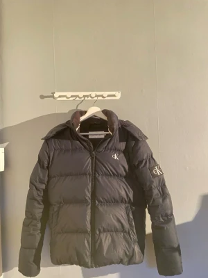 Calvin Klein Jacka - Säljer en svart pufferjacka från Calvin Klein Jeans med broderad CK-logga på bröstet och ärmen. Jackan har huva, dragkedja framtill och quiltad design. Perfekt för kalla dagar och ger en clean, modern look. //Skick 8/10// //Färg-Marinblå// // Storlek S//